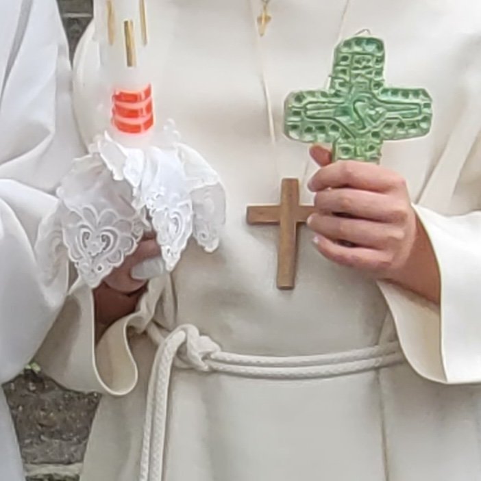 Eine Kinderhand, die ein grünes Kreuz in der Hand hält. Eine zweite Kinderhand hält eine Kerze. Zu sehen ist ein helles Erstkommunionskleid mit Kordel und Holzkreuz.